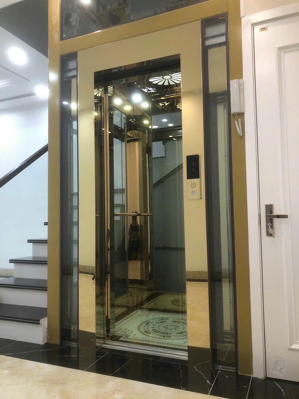Thang máy gia đình cửa kính khung vàng sang trọng lắp đặt cạnh cầu thang trong nhà